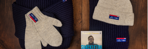 National Maritime Museum X Arthur Beale products against a wooden background. Products left to right: folded navy submariner sweater, grey woollen mittens folded on top of the submariner, Shackleton book by Penguin, navy woolen scarf, grey woollen beanie.