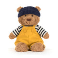Teddy bear wearing a navy hat, striped shirt, and yellow dungarees