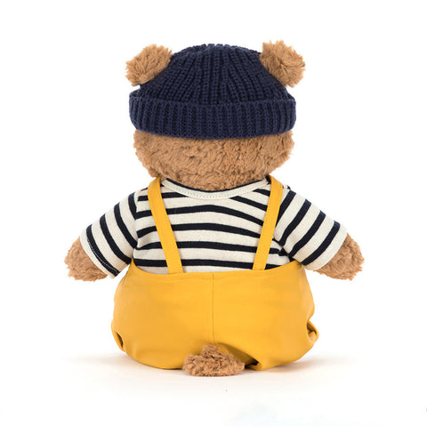 Teddy bear wearing a navy hat, striped shirt, and yellow overalls on a white background