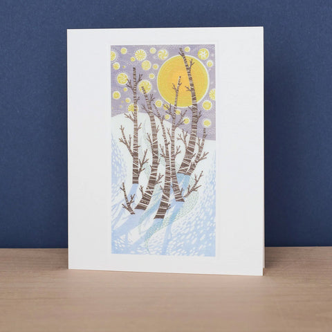 A greeting card featuring a stylized winter forest illustration with birch trees, a yellow sun, and a lavender sky, standing on a light wood surface against a dark blue background.