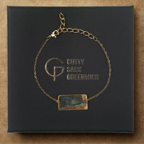 Gold bracelet with a rectangular greenish piece of brass on a black box with 'Cutty Sark Greenwich' branding