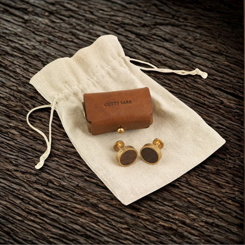 Gold cufflinks with a wooden box labeled 'Cutty Sark' on a textured brown surface.
