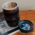 A novelty travel mug shaped like a Canon zoom lens, filled with coffee and placed on a wooden table next to its lens cap and a camera manual.