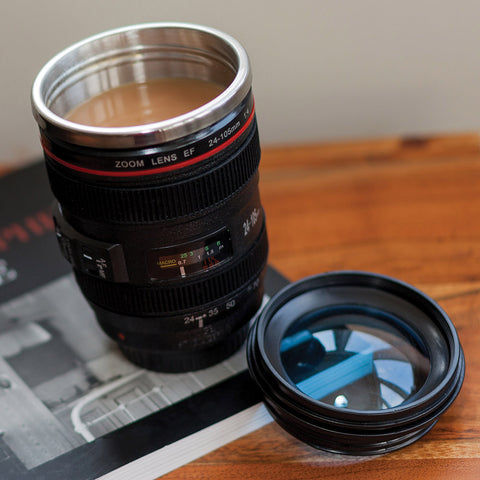 A novelty travel mug shaped like a Canon zoom lens, filled with coffee and placed on a wooden table next to its lens cap and a camera manual.
