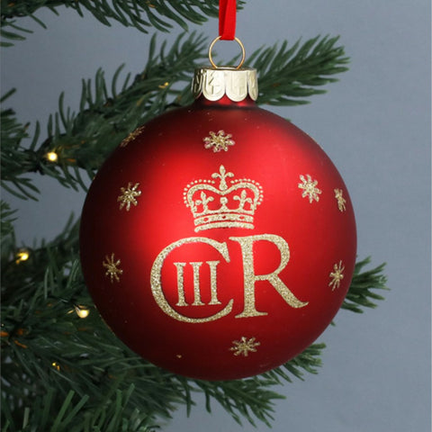 A red Christmas bauble ornament with a gold crown and the initials 'CR' in glittery gold, hanging from a green Christmas tree branch with warm string lights against a gray background.