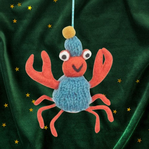 Decorative crab ornament with a blue sweater and yellow pom-pom on a green background with gold stars.