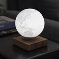 A glowing 3D spherical moon map lamp floating above a square wooden base, placed on a dark table in a dimly lit indoor setting.