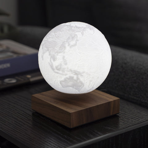 A glowing 3D spherical moon map lamp floating above a square wooden base, placed on a dark table in a dimly lit indoor setting.