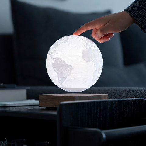 A glowing globe night light floating above a wooden base on a dark table, with a person's hand touching the top of the sphere in a dim living room setting.