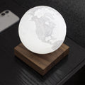 A glowing 3D moon-phase style globe lamp resting on a square wooden base, photographed on a dark tabletop with a smartphone partially visible in the background.