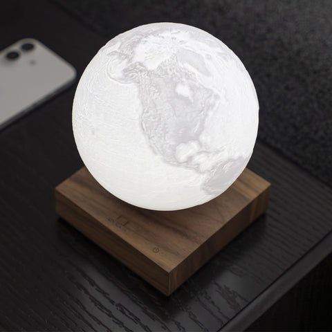 A glowing 3D moon-phase style globe lamp resting on a square wooden base, photographed on a dark tabletop with a smartphone partially visible in the background.