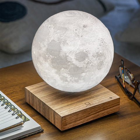 A 3D-printed moon lamp with detailed craters and textures floating above a wooden base, set on a wooden table next to a notebook and eyeglasses.