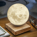 A 3D-printed moon lamp floating above a wooden base on a wooden table, with a notebook, eyeglasses, and a blurred home background.