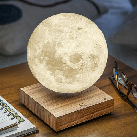 A 3D-printed moon lamp floating above a wooden base on a wooden table, with a notebook, eyeglasses, and a blurred home background.
