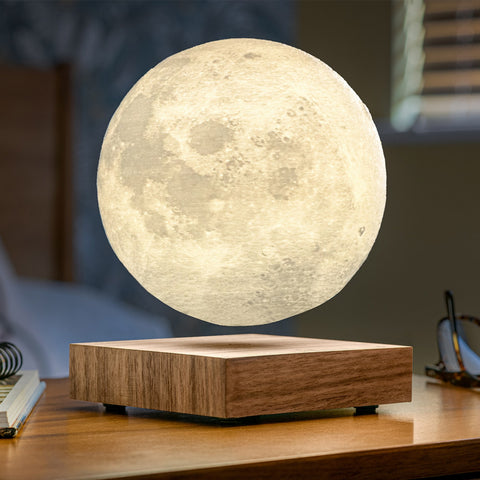 A glowing moon-shaped night light floating above a square wooden base on a wooden table, with a softly blurred bedroom background.