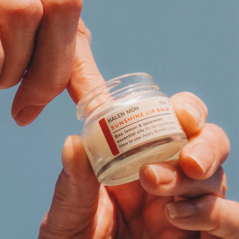 A close-up of hands holding a small clear jar of HALEN MÖN Sunshine Lip Balm against a solid blue sky background.