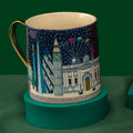 A ceramic mug with a gold handle and a winter cityscape illustration featuring Big Ben, the London Eye, and modern skyscrapers under a dark blue night sky, displayed on a teal circular stand against a dark green background.
