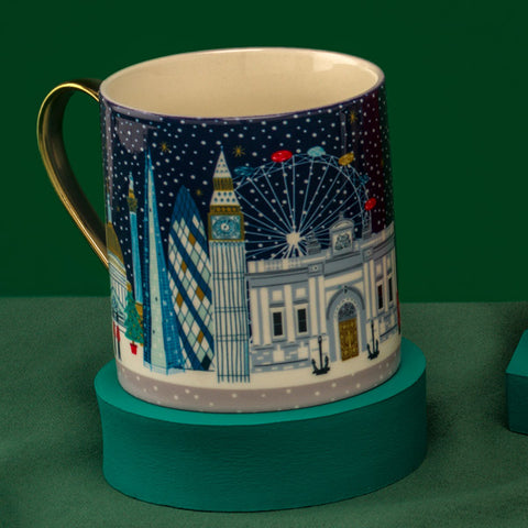 A ceramic mug with a gold handle and a winter cityscape illustration featuring Big Ben, the London Eye, and modern skyscrapers under a dark blue night sky, displayed on a teal circular stand against a dark green background.