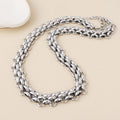 A silver-toned multi-strand chain necklace with a textured, woven appearance, arranged in a loop on a light beige background with a small white decorative object in the top-left corner.
