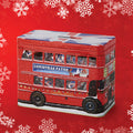 A red double-decker bus-shaped Christmas tin with snowflake details, featuring a 'Christmas Patter' advertisement on the side, set against a red background with white snowflakes.