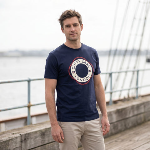 Man wearing a navy blue t-shirt with a logo standing on a wooden deck by a body of water.