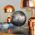 Decorative moon sphere on a stand with two copper lamps in the background.