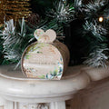 A round scented candle in a decorative paper sleeve with a heart-shaped handle, placed on a white mantel in front of a decorated Christmas tree.