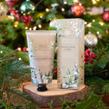 A tube and box of Under The Mistletoe Hand Cream placed on a wooden slice in front of a decorated Christmas tree with lights and holly.