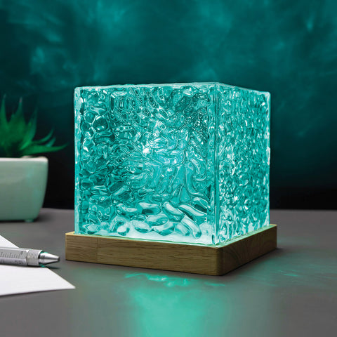 A square glass cube lamp with a textured, hammered surface glowing in a bright teal-green light, resting on a wooden base on a desk.