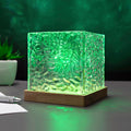 A square glass cube lamp with a textured, hammered surface glowing green, resting on a wooden base on a desk.
