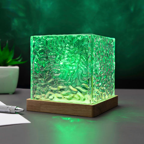 A square glass cube lamp with a textured, hammered surface glowing green, resting on a wooden base on a desk.