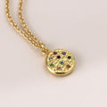 Gold necklace with a round pendant featuring colorful gemstones on a light pink background