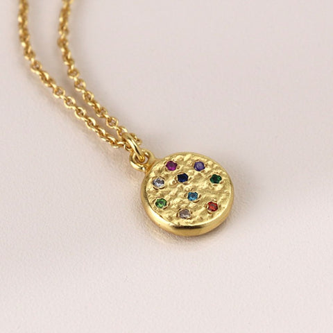 Gold necklace with a round pendant featuring colorful gemstones on a light pink background