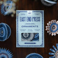 Pack of East End Press ornaments on a wooden surface with decorative paper ornaments around.
