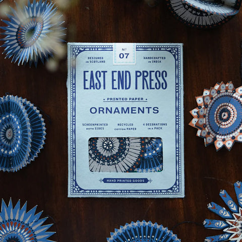 Pack of East End Press ornaments on a wooden surface with decorative paper ornaments around.