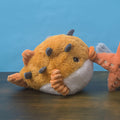 A plush toy fish with a fuzzy orange-brown body, white belly, and small gray spikes, resting on a dark wooden surface against a blue background. Part of another orange plush toy is visible at the right edge.