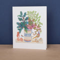 A white art print or greeting card standing upright on a light wood surface against a dark blue wall. The printed illustration shows a vase filled with green and pink botanical elements, with small birds and decorative motifs around it.