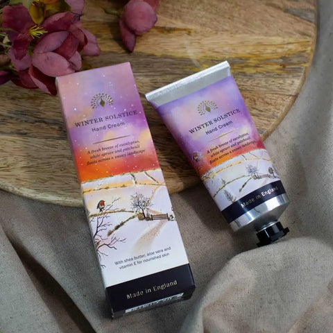 Winter Solstice Hand Cream tube and matching box on a rustic wooden board with fabric and dried flowers in the background. The packaging features a winter landscape design with a purple sky, orange sunset gradient, and snowy branches.