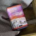 A tin of Winter Solstice Hand & Body Soap by The English Soap Company, styled on a textured gray blanket next to a wooden surface.