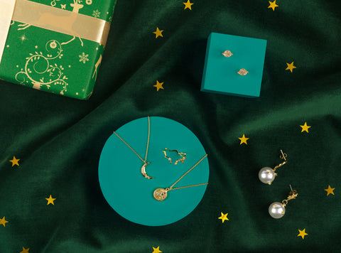 Gold crescent moon necklace and gold jewelled pendant necklace on green background, surrounded by constellation gold ring, gold Saturn earrings and gold pearl drop earrings.