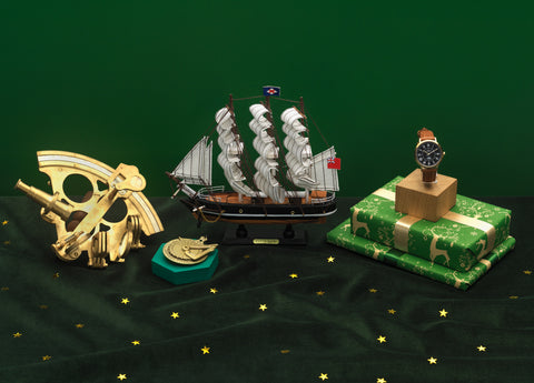 Gold sextant replica, golden replica sundial, Cutty Sark model ship and The Camden Watch Company Cutty Sark Royal Museums Greenwich watch arranged on a green starry setting with wrapped Christmas gifts