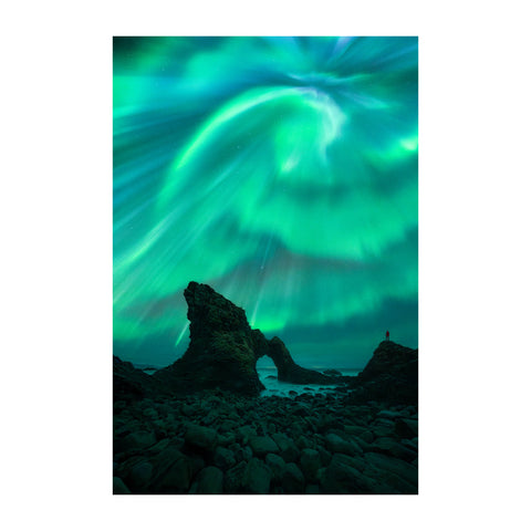 A vertical photographic print of the northern lights over a rocky coastal landscape, with glowing green and teal aurora sweeping across the night sky above dark rock formations.