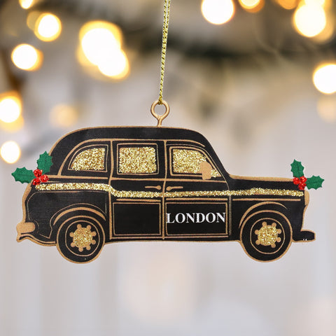 A black London cab Christmas ornament with gold glitter details, green holly leaves, and red berries, hanging against a blurred background of warm bokeh lights.
