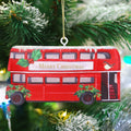 A red double-decker bus Christmas ornament hanging from a gold string on a decorated Christmas tree. The ornament features 'Merry Christmas' in gold lettering on the front, holly leaves, red berries, a small bird, and snow-dusted details.
