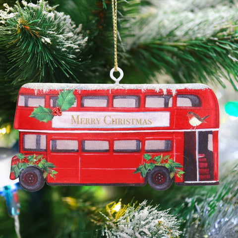 A red double-decker bus Christmas ornament hanging from a gold string on a decorated Christmas tree. The ornament features 'Merry Christmas' in gold lettering on the front, holly leaves, red berries, a small bird, and snow-dusted details.