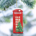 A red telephone booth Christmas ornament hanging from a gold string, decorated with a small green Christmas tree and ornaments, set against a blurred snowy pine background.