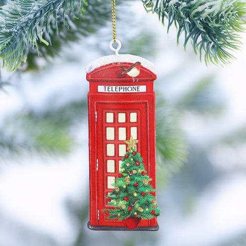 A red telephone booth Christmas ornament hanging from a gold string, decorated with a small green Christmas tree and ornaments, set against a blurred snowy pine background.