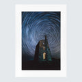 A vertical photographic art print featuring a stone tower under a swirling star trail night sky, presented with a wide white border 