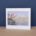 A white matted art print card featuring a stylized illustration of a robin bird perched on a branch with red berries against a soft lavender and blue landscape, displayed on a light wood surface against a dark blue background.