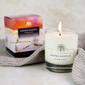 A lit white soy wax candle in a clear glass jar sits on a light surface beside its colorful product box. The candle label reads Winter Solstice by The Solar Company, and a knitted gray textile is draped around the base.